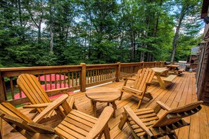 Terraza de madera con sillas Adirondack y una mesa, rodeada de árboles.