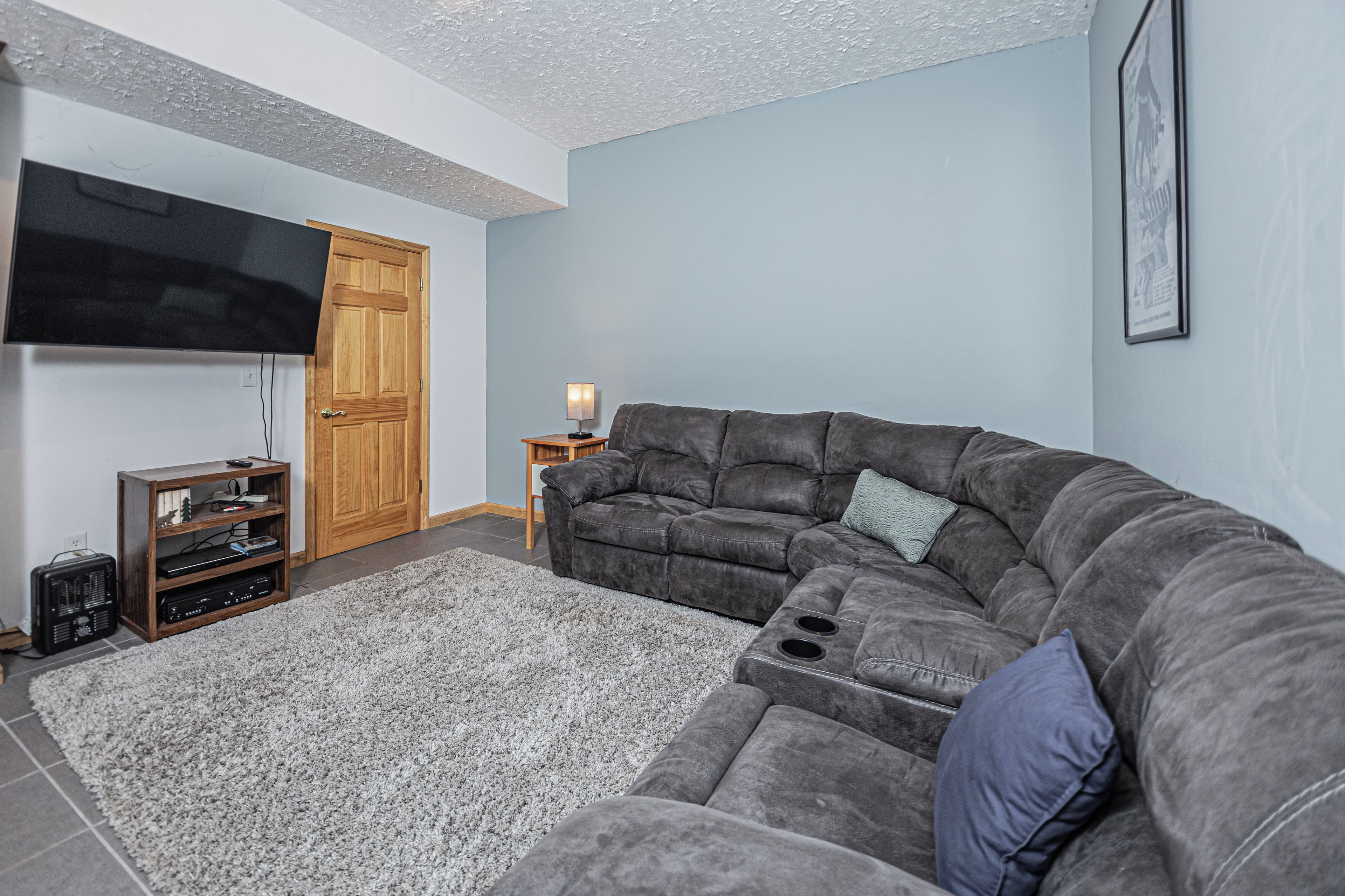 Ein Wohnzimmer mit einem Ecksofa, einem flachbildfernseher an der Wand, einer Holzkommode und einem kleinen Tisch mit einer Lampe.