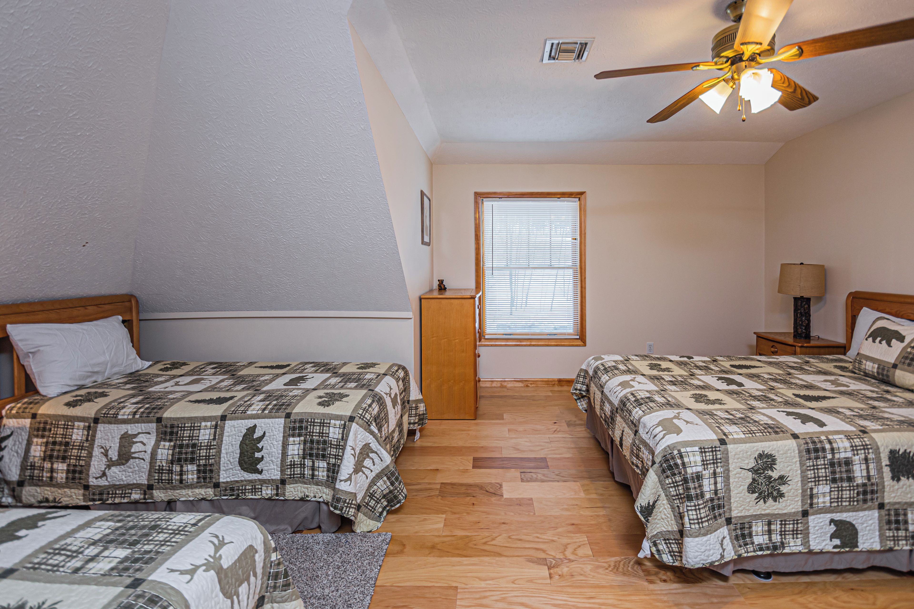 A bedroom with two beds, wooden flooring, a ceiling fan, a window with blinds, and a nightstand with a lamp.
