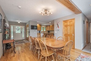 A spacious dining area with a wooden table and chairs, a kitchen in the background, and a wooden door with a poster.