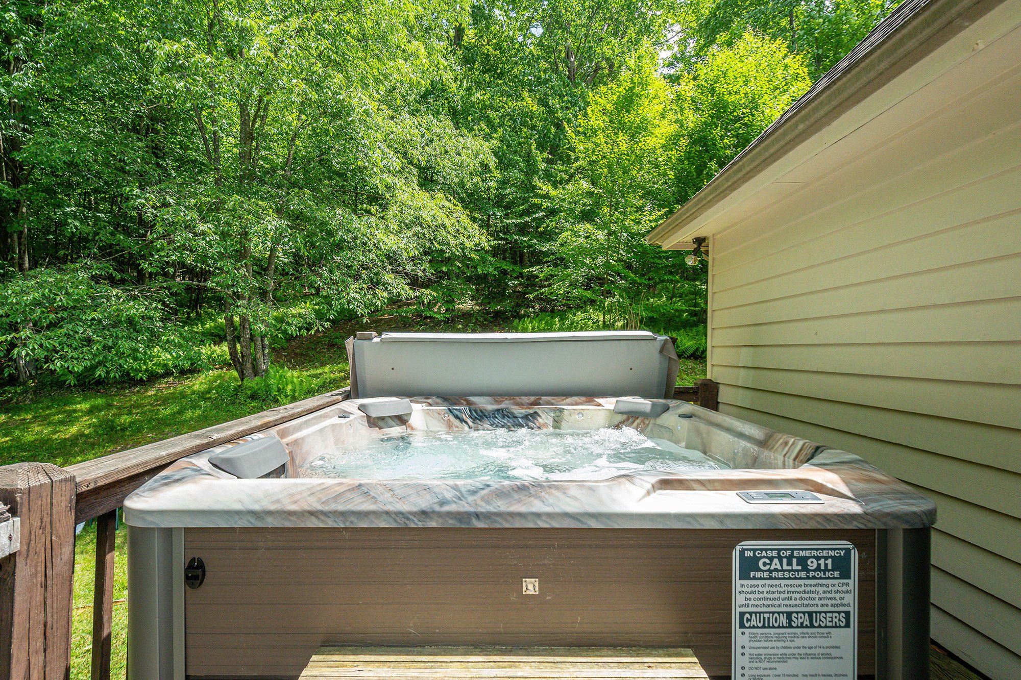 Un bain à remous installé dans un jardin, avec une terrasse en bois et un panneau indiquant les coordonnées des personnes à contacter en cas d’urgence.