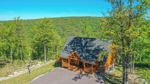 A log cabin with a porch and a covered patio, surrounded by trees and a mountain in the background. - Moonlight Ridge by Taylor Made Deep Creek (McHenry)