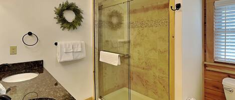 A bathroom with a glass-enclosed shower, two sinks, a toilet, and a window with blinds.