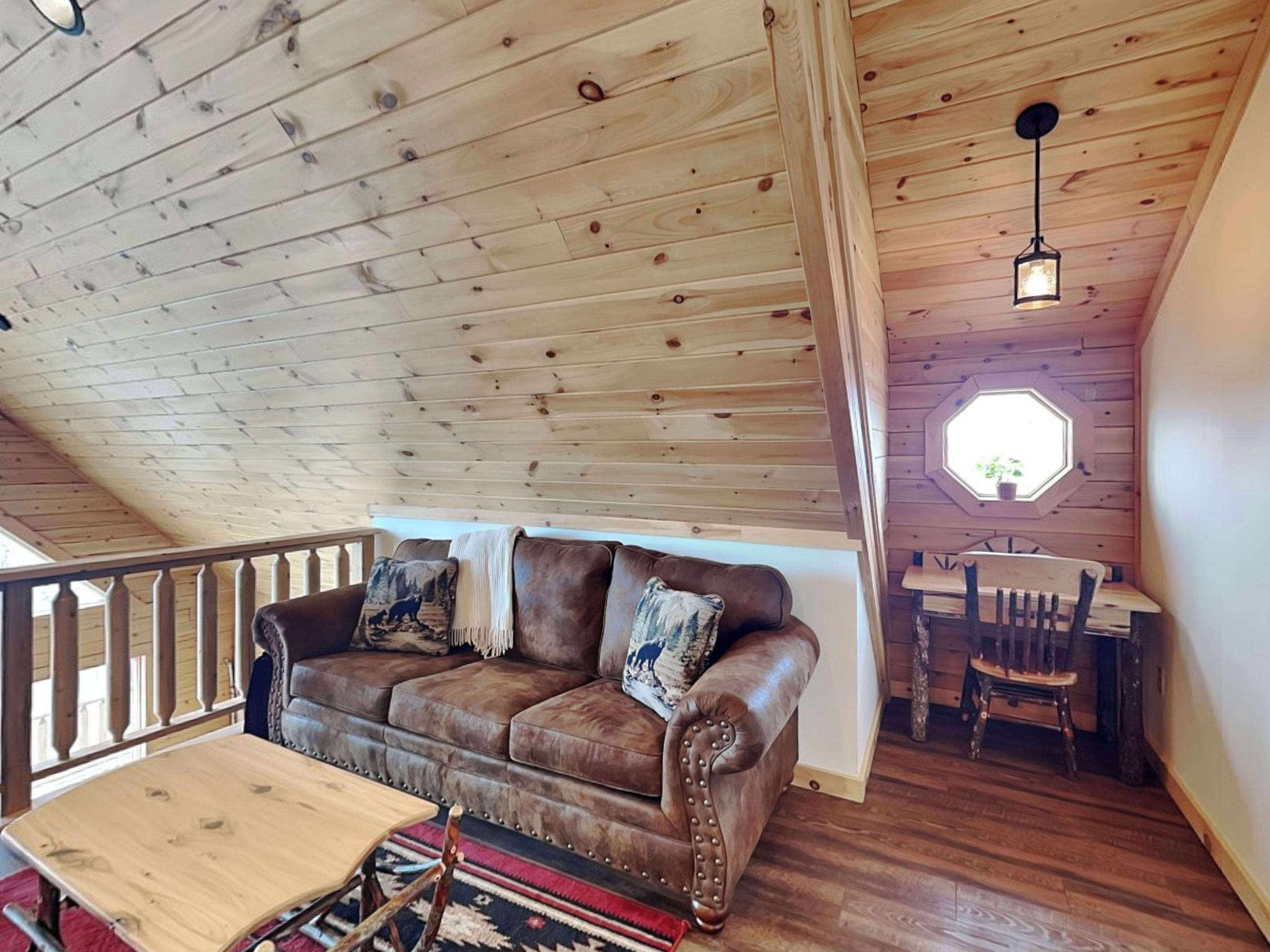 A loft-style room with a wooden ceiling, a brown leather sofa with decorative pillows, a wooden dining table with chairs, and a small window with a plant.