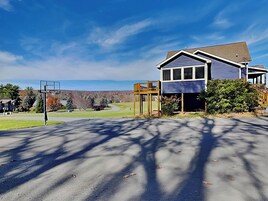 A residential house with a basketball court in the foreground and a clear view of a hilly landscape in the background.