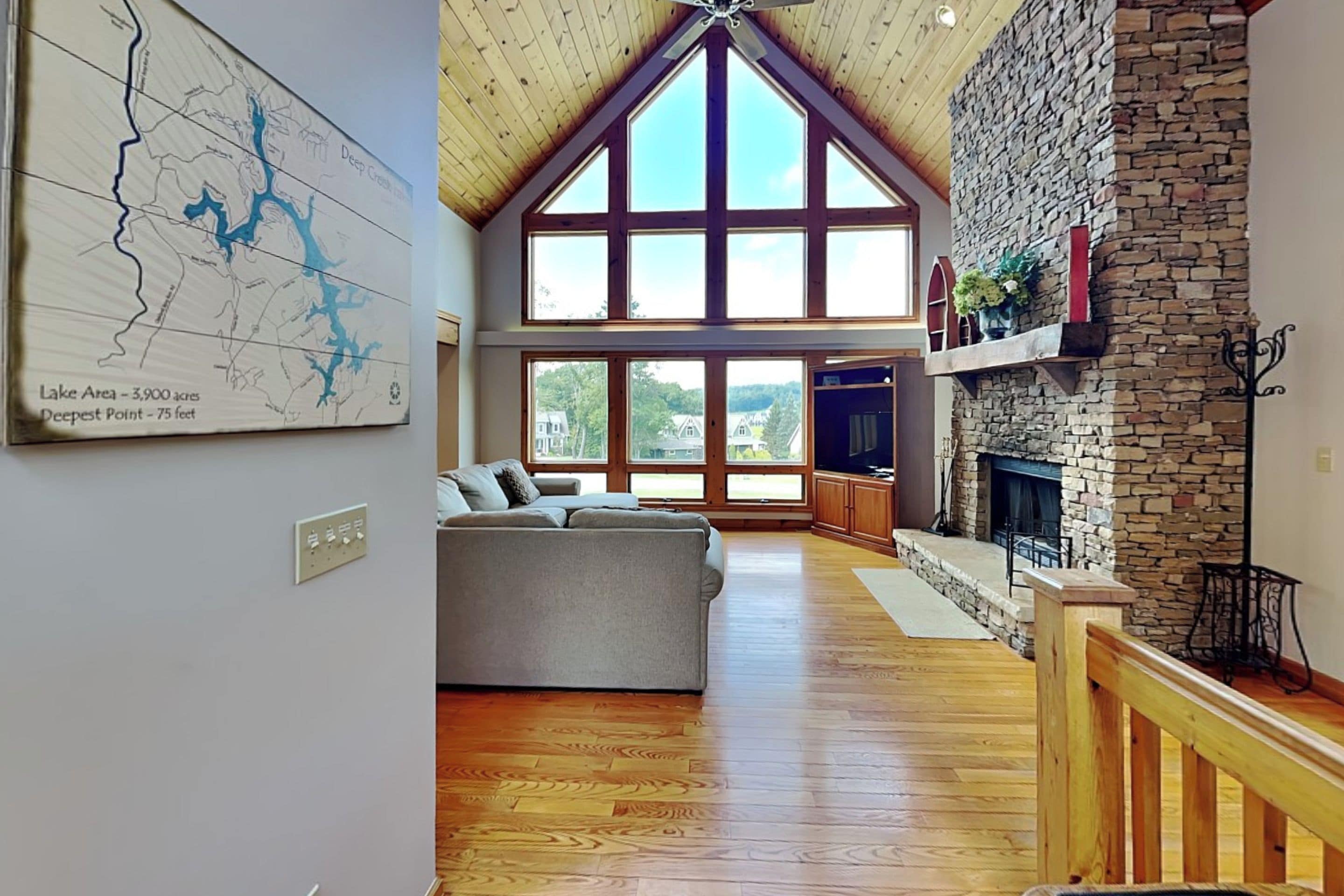 A spacious living room with a high ceiling, large windows, a stone fireplace, and a map on the wall.