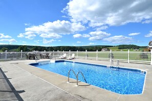 Uma piscina retangular com escada de metal, rodeada por uma cerca branca e um deck com cadeiras.