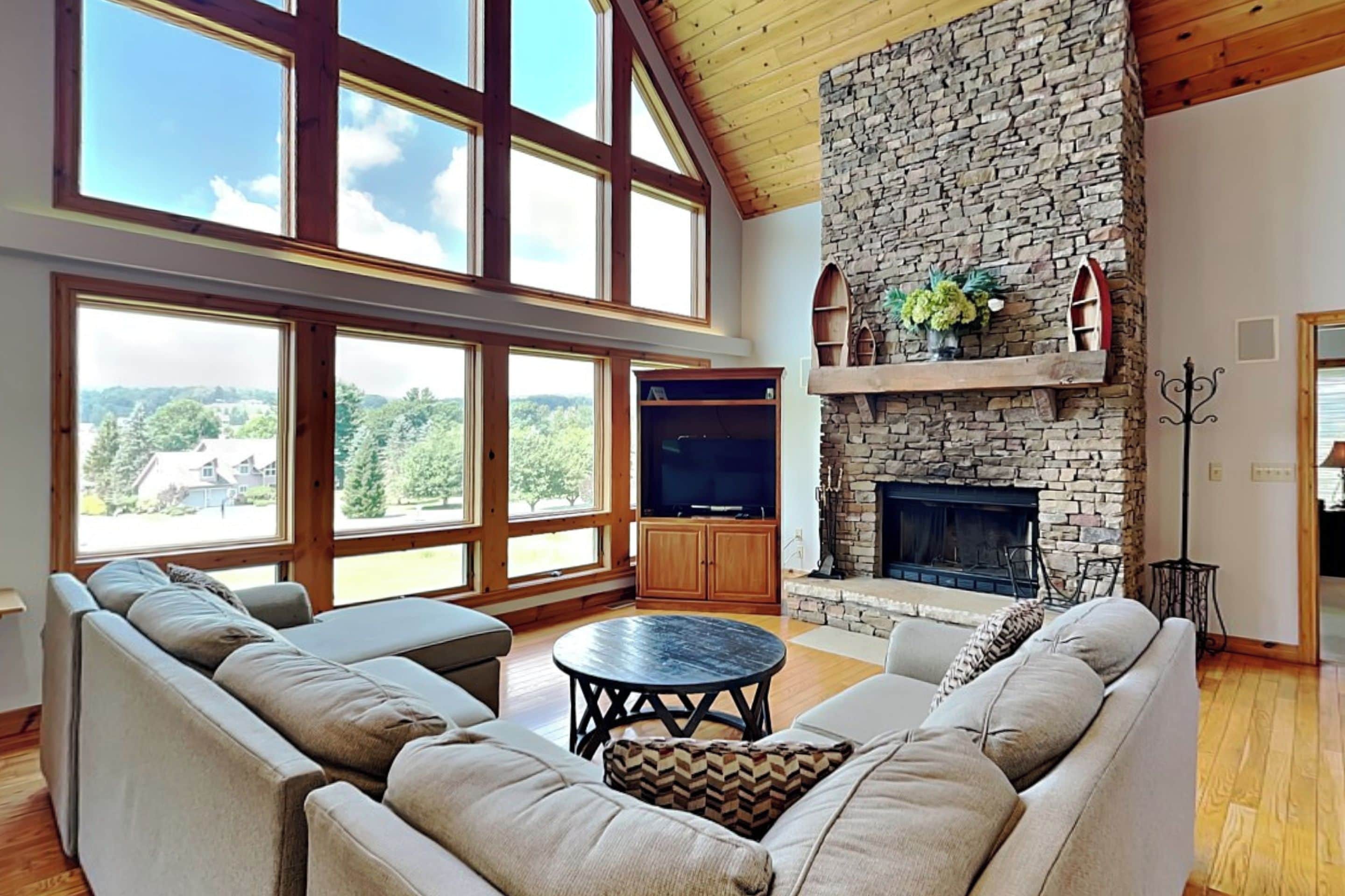 A spacious living room with a stone fireplace, large windows, and a high ceiling.