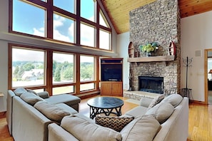 A spacious living room with a stone fireplace, large windows, and a high ceiling.