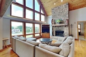 A spacious living room with a stone fireplace, large windows, and a high ceiling.