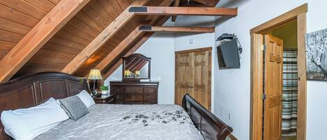 A bedroom with a sloped ceiling, wooden beams, a bed with a grey and white bedspread, a nightstand with a lamp, a dresser, a mirror, and a door leading to another room.