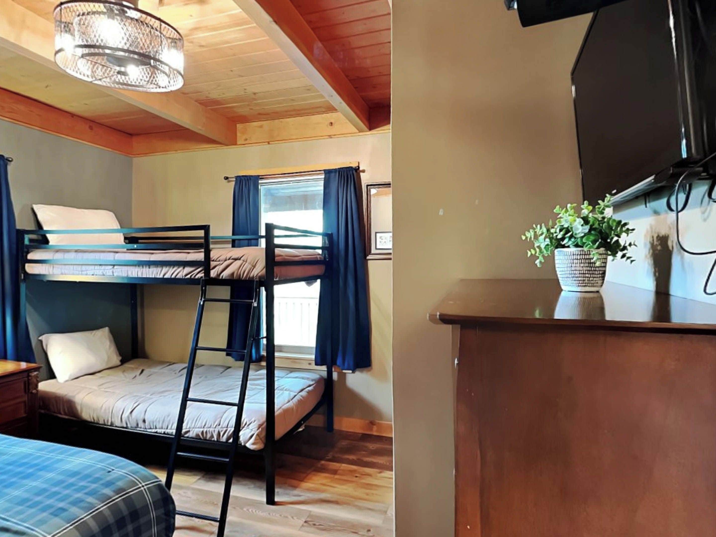 A bunk bed room with a wooden ceiling, a desk with a plant, and a window with curtains.