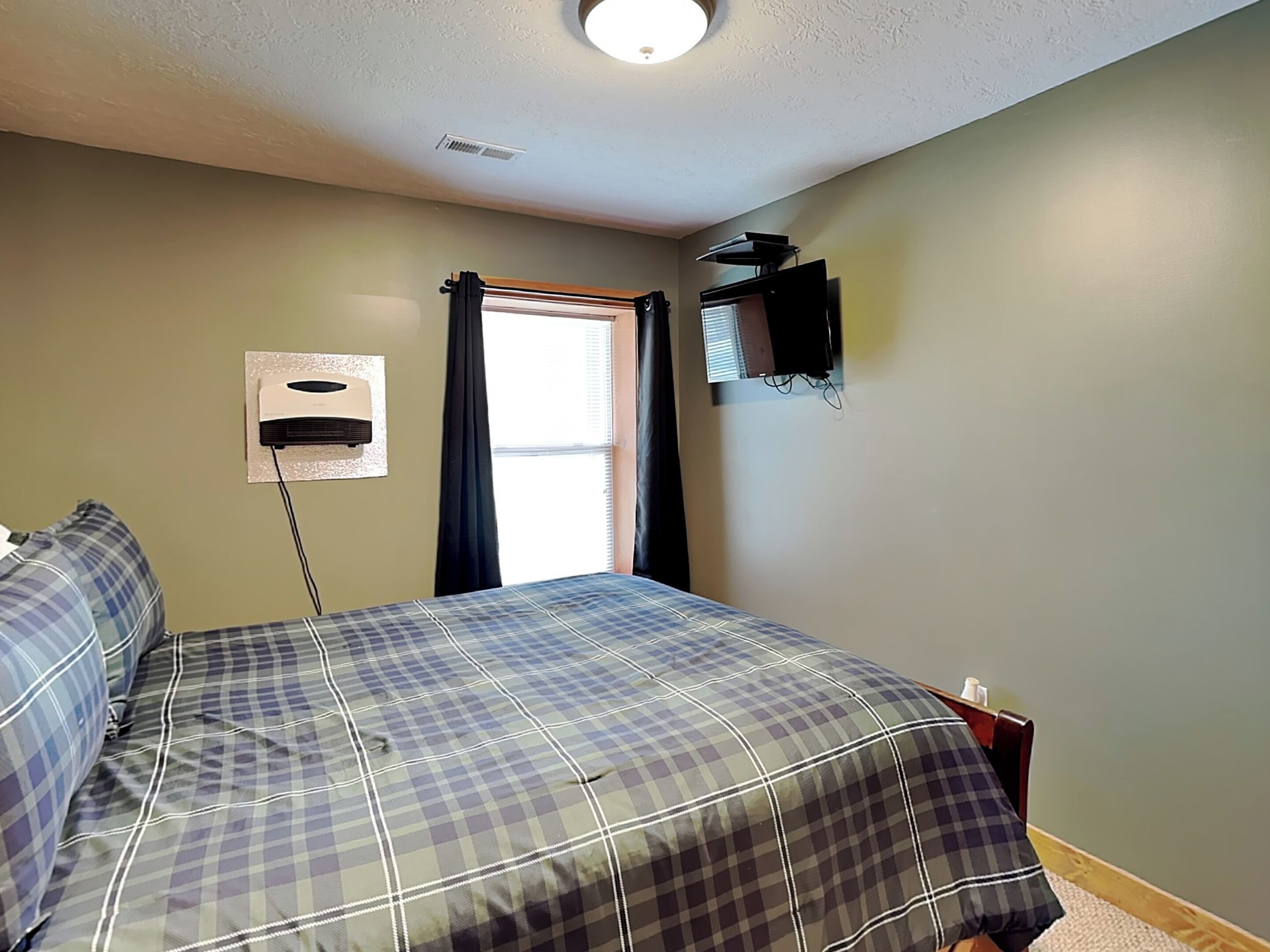 A bedroom with a bed, a window with curtains, a wall-mounted air conditioner, and a television on the wall.