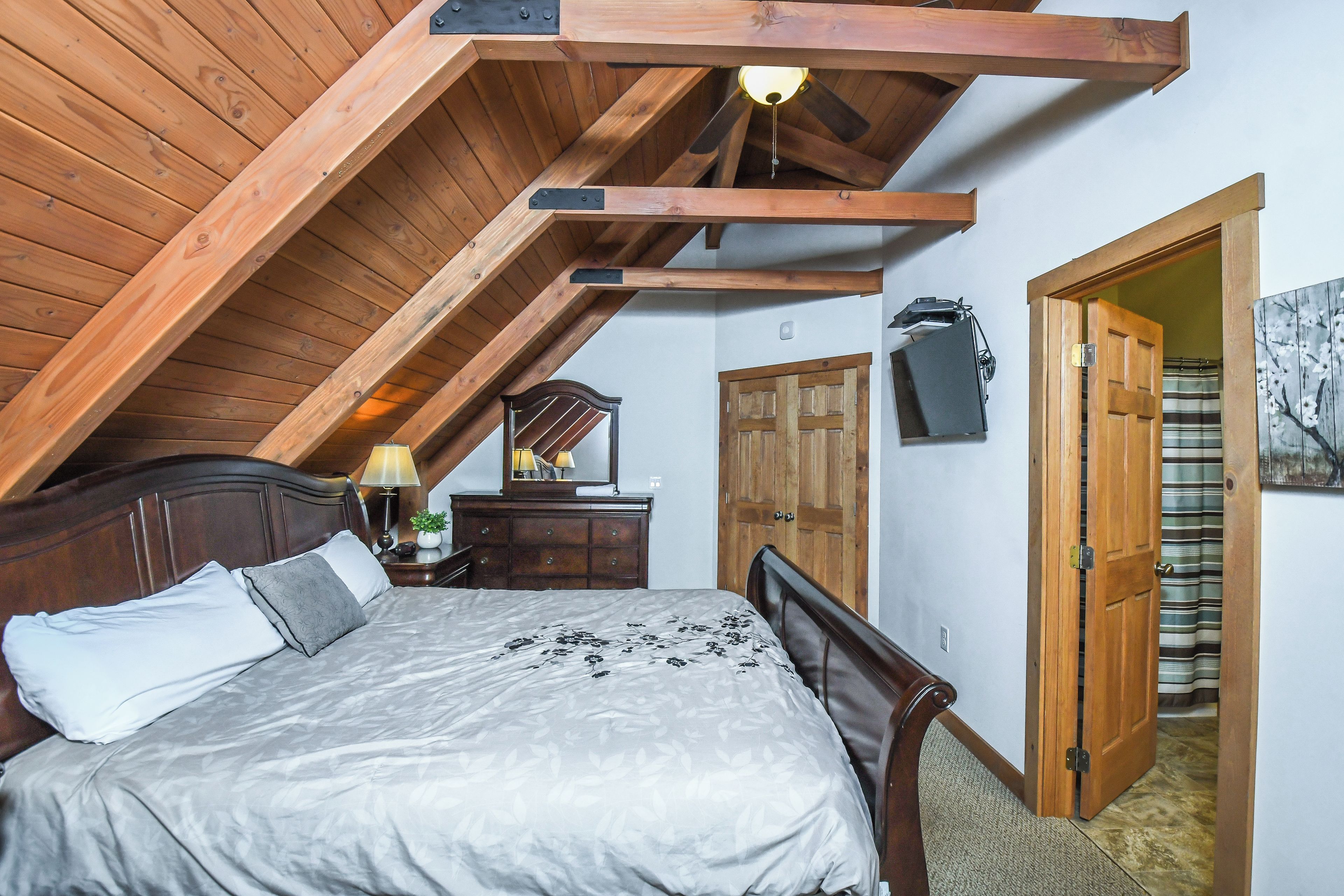 Une chambre avec un plafond en pente, des poutres en bois, un lit recouvert d’une couvre-lit grise et blanche, une table de chevet avec une lampe, une commode, un miroir et une porte donnant sur une autre pièce.