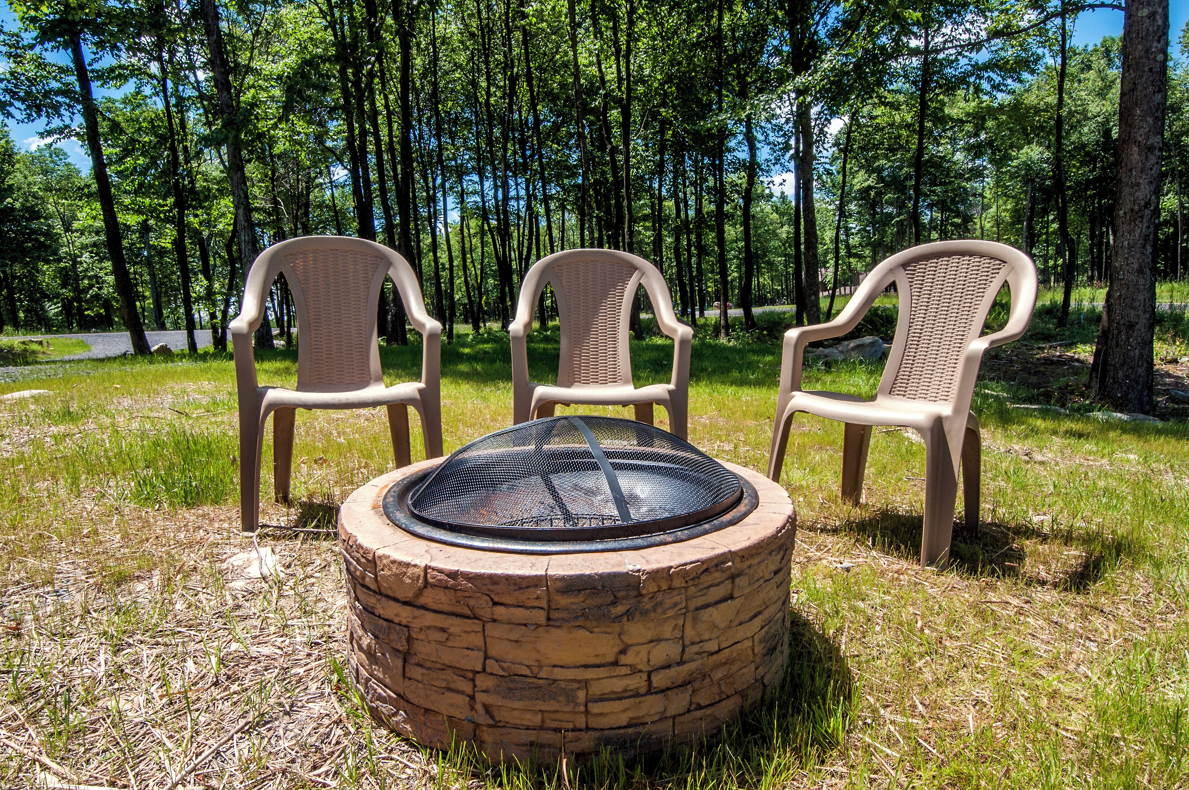 Three wicker chairs and a stone fire pit in a grassy area surrounded by trees.