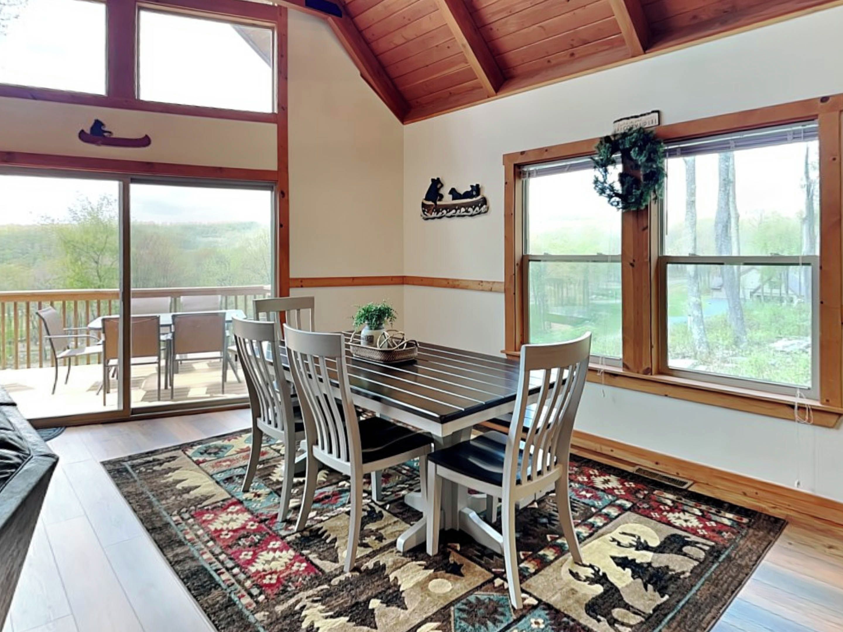 A dining area with a table and chairs, a patterned rug, and large windows with a view of trees.