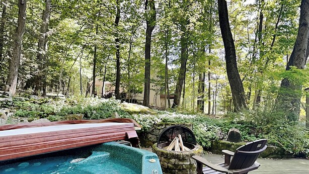 A hot tub surrounded by a wooden deck, a stone fire pit, and a lounge chair in a forested area. - Mountain Escape by Taylor Made Deep Creek (McHenry)