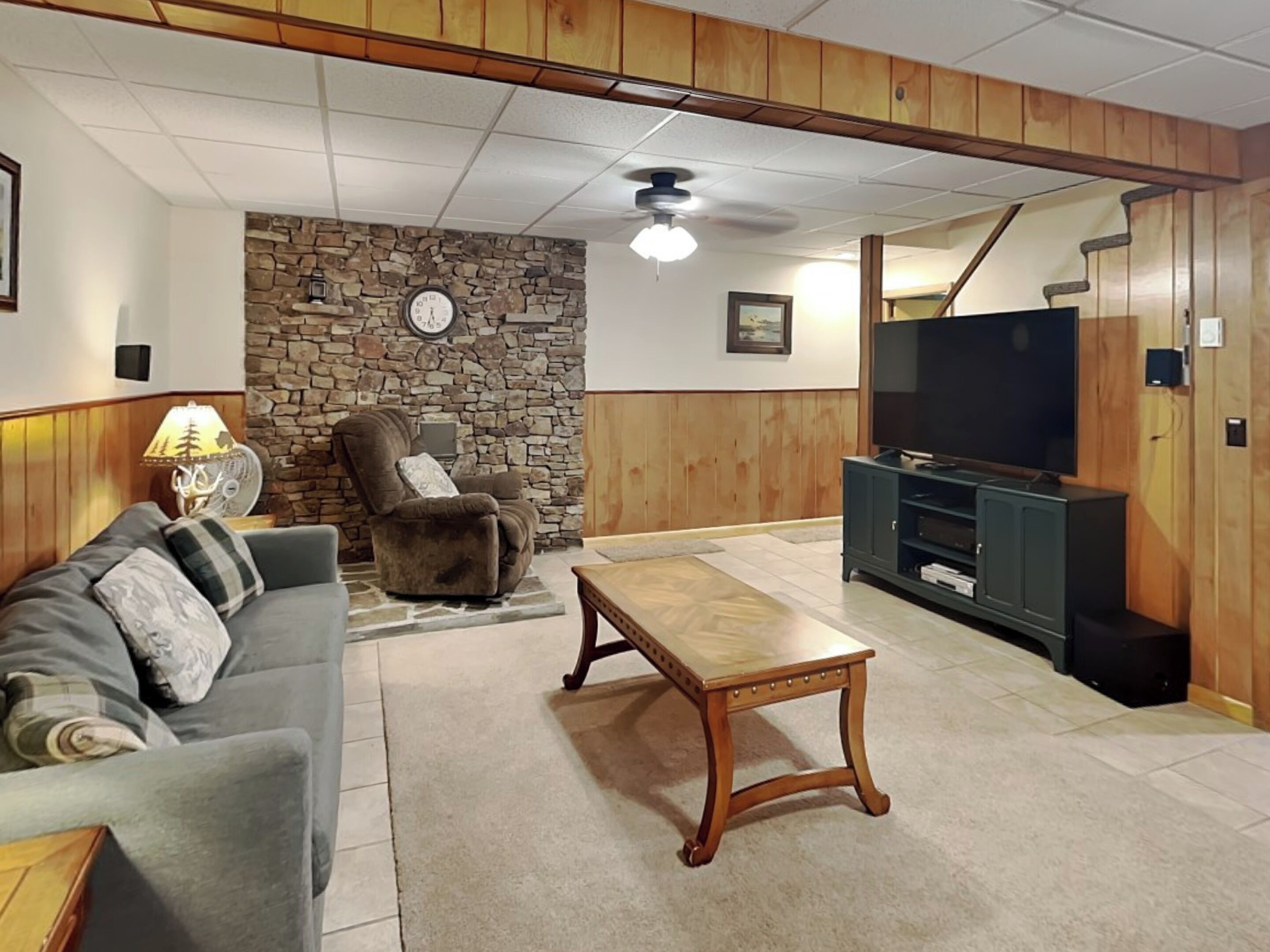 Ein Wohnzimmer mit einer Steinwand, einer Holzbalkendecke, einem grauen Sofa, einem Holztisch, einem Liegestuhl, einem TV-Schrank und einem Deckenventilator.