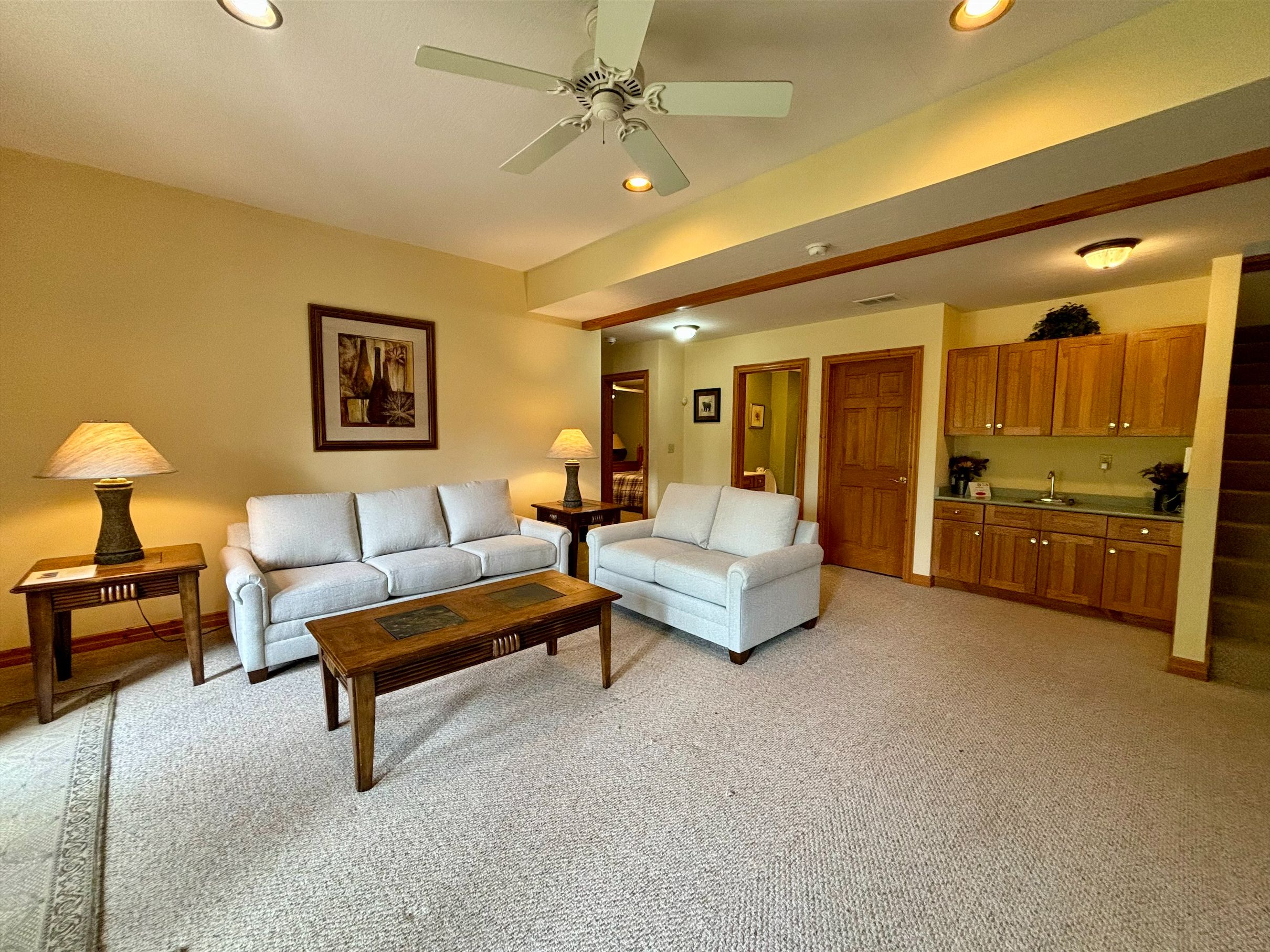 A living room with two sofas, a coffee table, and a ceiling fan. There is a kitchen area visible through an open doorway.