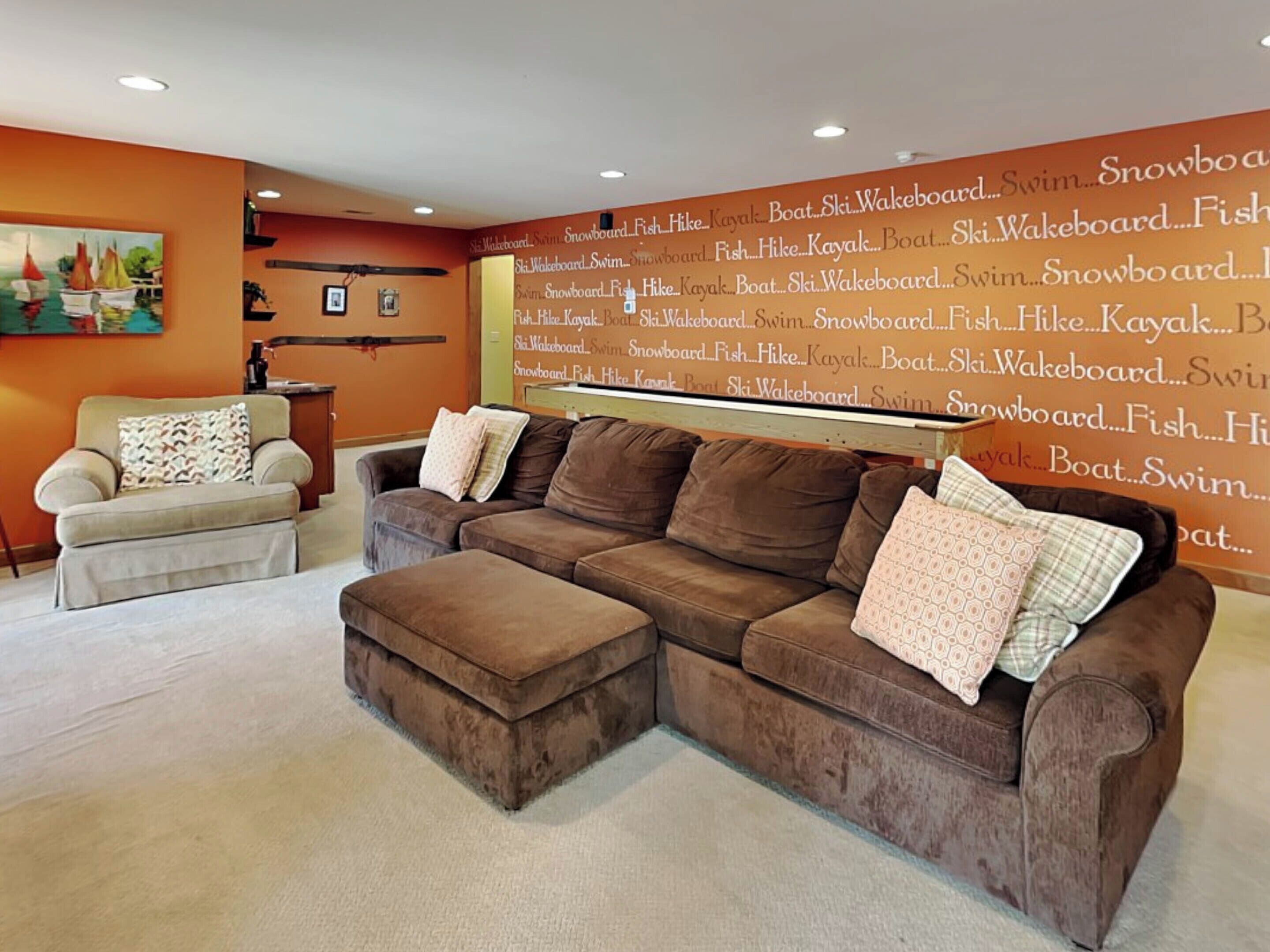 A living room with a brown sectional sofa, a beige armchair, and a coffee table. A large wall mural lists various water sports activities.