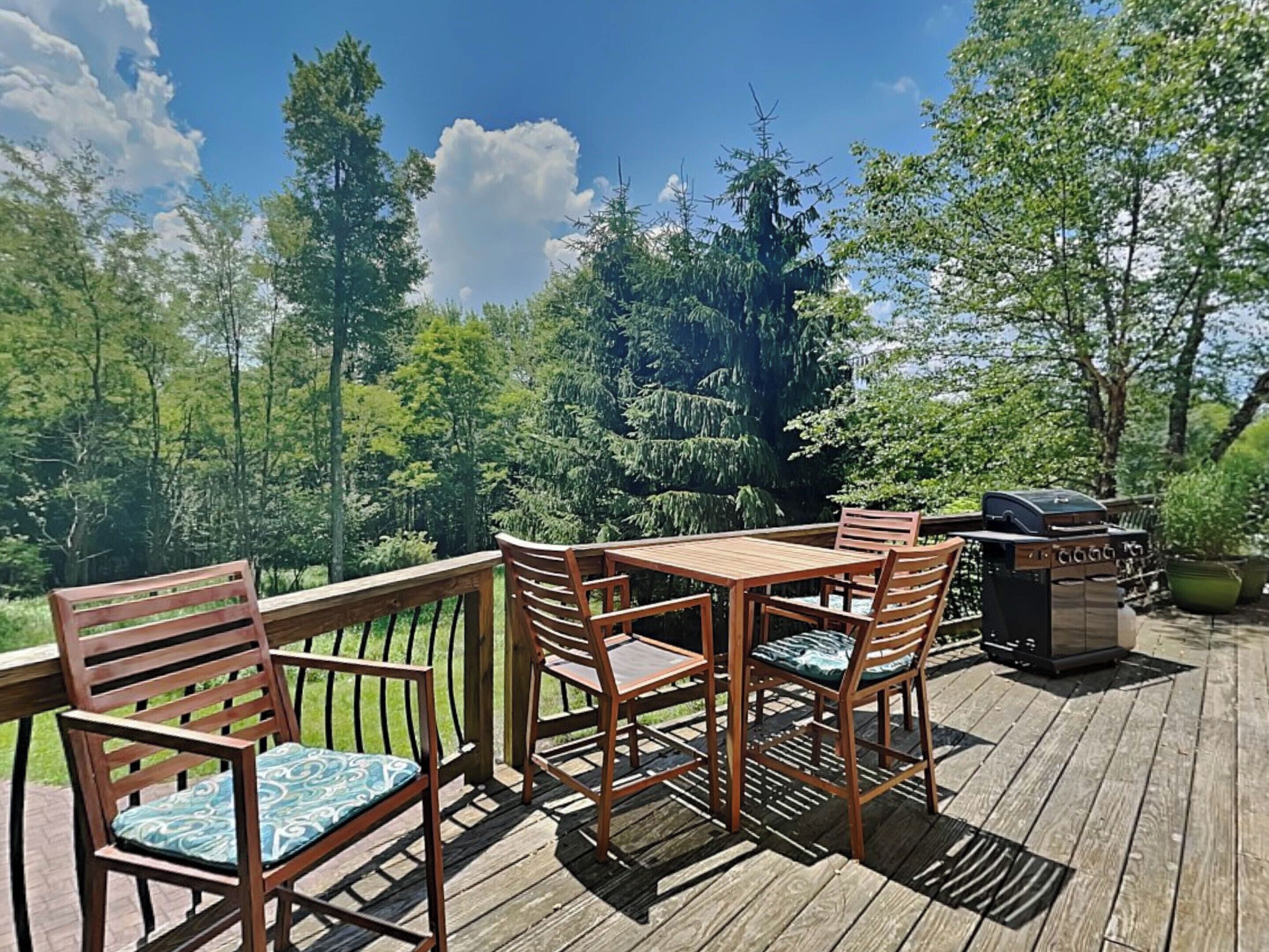 Una terraza de madera con mesa y sillas, una parrilla y vistas a un área boscosa.