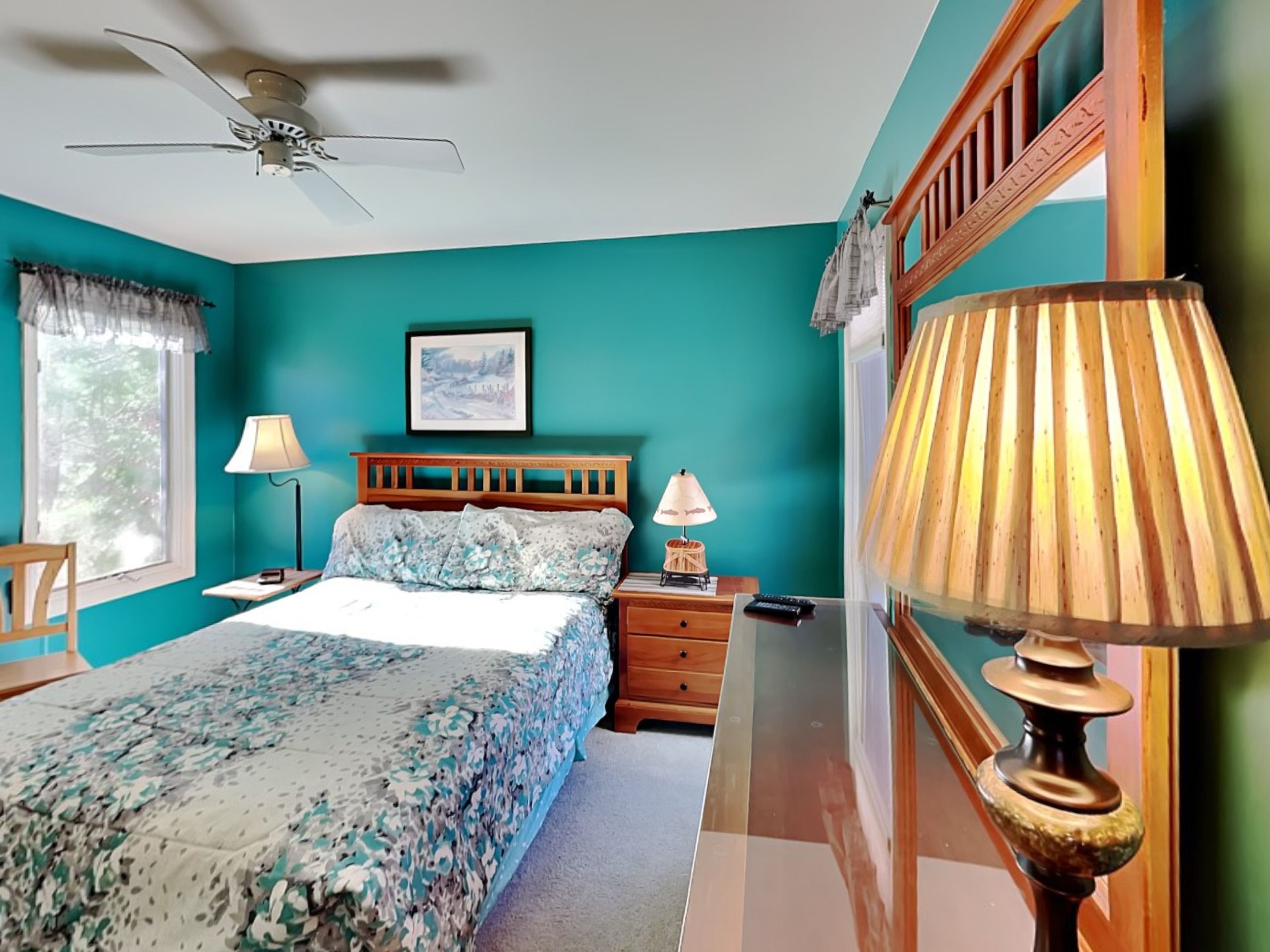 A bedroom with teal walls, a bed with floral bedding, a ceiling fan, two bedside lamps, a framed picture on the wall, and a wooden nightstand.