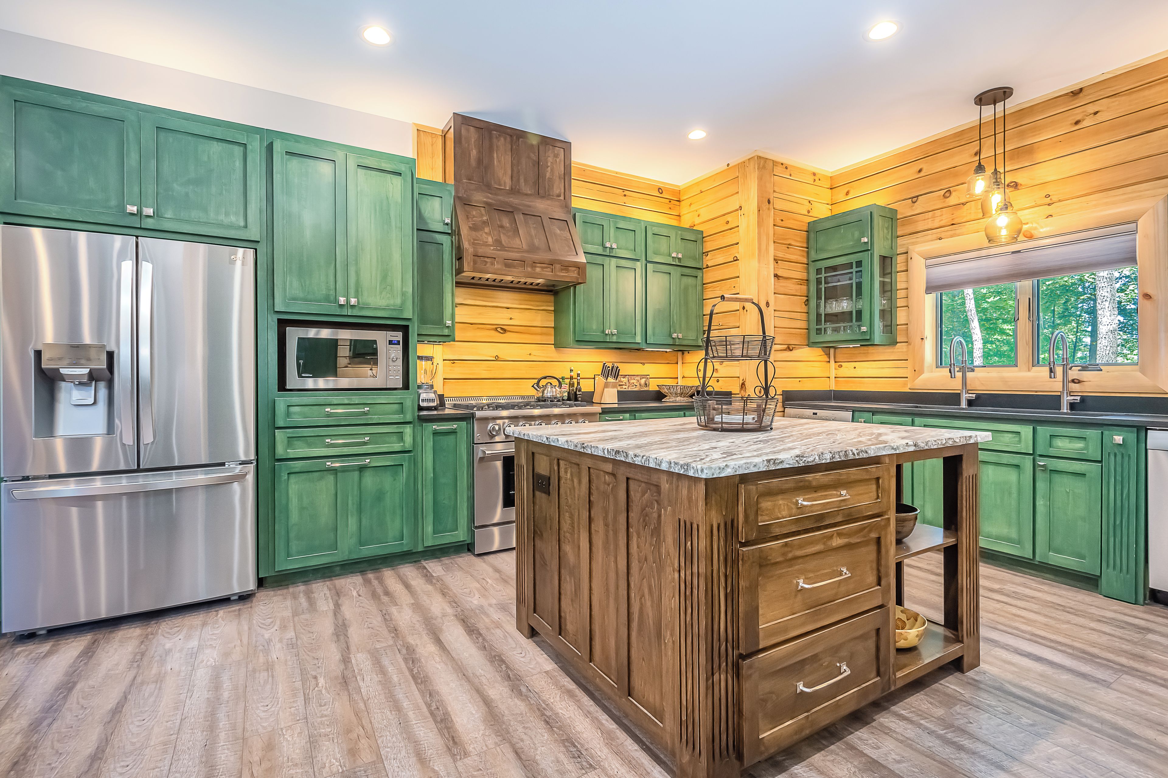 Una cocina con armarios verdes, un refrigerador de acero inoxidable, una isla de madera y una ventana con vista a árboles.