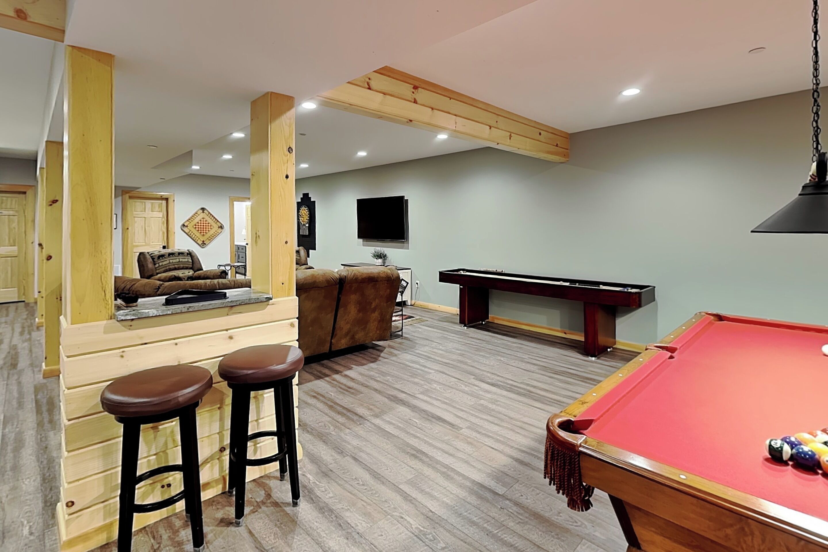 A pool table, bar stools, a sofa, and a television in a room with wooden beams and a light-colored floor.