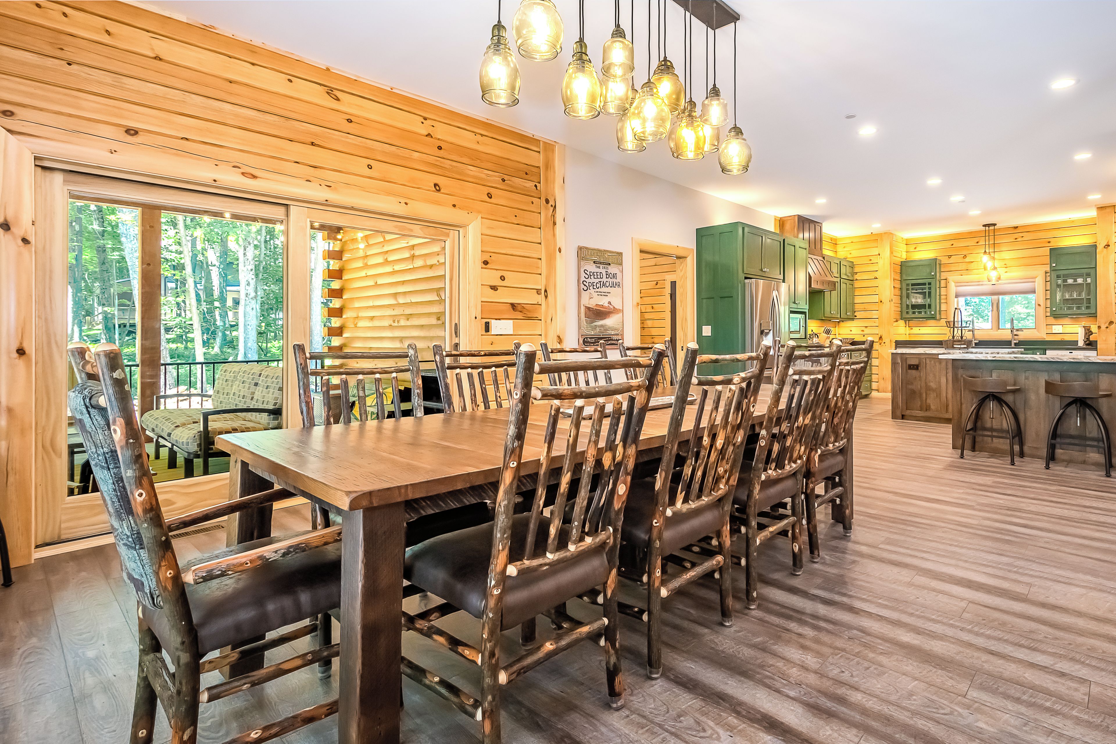 A wooden-paneled dining area with a long table and chairs, a kitchen with green cabinets, and large windows.