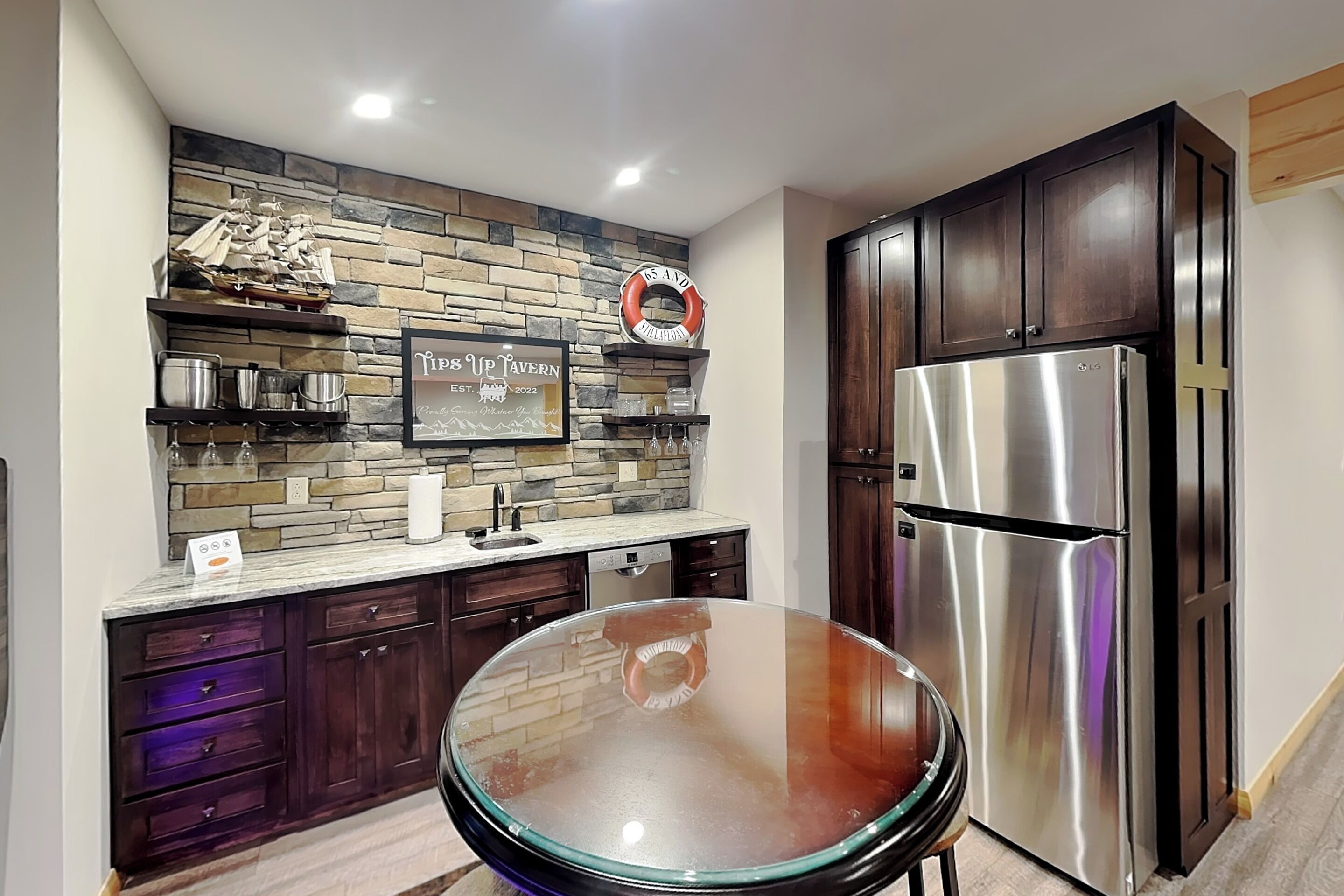 A kitchen with dark wooden cabinets, a stainless steel refrigerator, a stone backsplash, and a glass-top table.