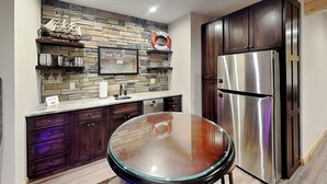 A kitchen with dark wooden cabinets, a stainless steel refrigerator, a stone backsplash, and a glass-top table. - Off the Deep End by Taylor Made Deep Creek (McHenry)