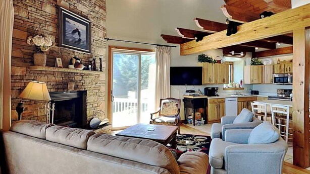 A living room with a stone fireplace, a sofa, a television, and a kitchen area with wooden beams. - On Eagle's Wings by Taylor Made Deep Creek (McHenry)