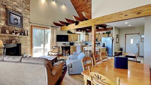 A spacious living area with a fireplace, a dining table with chairs, a kitchen in the background, and a large window.