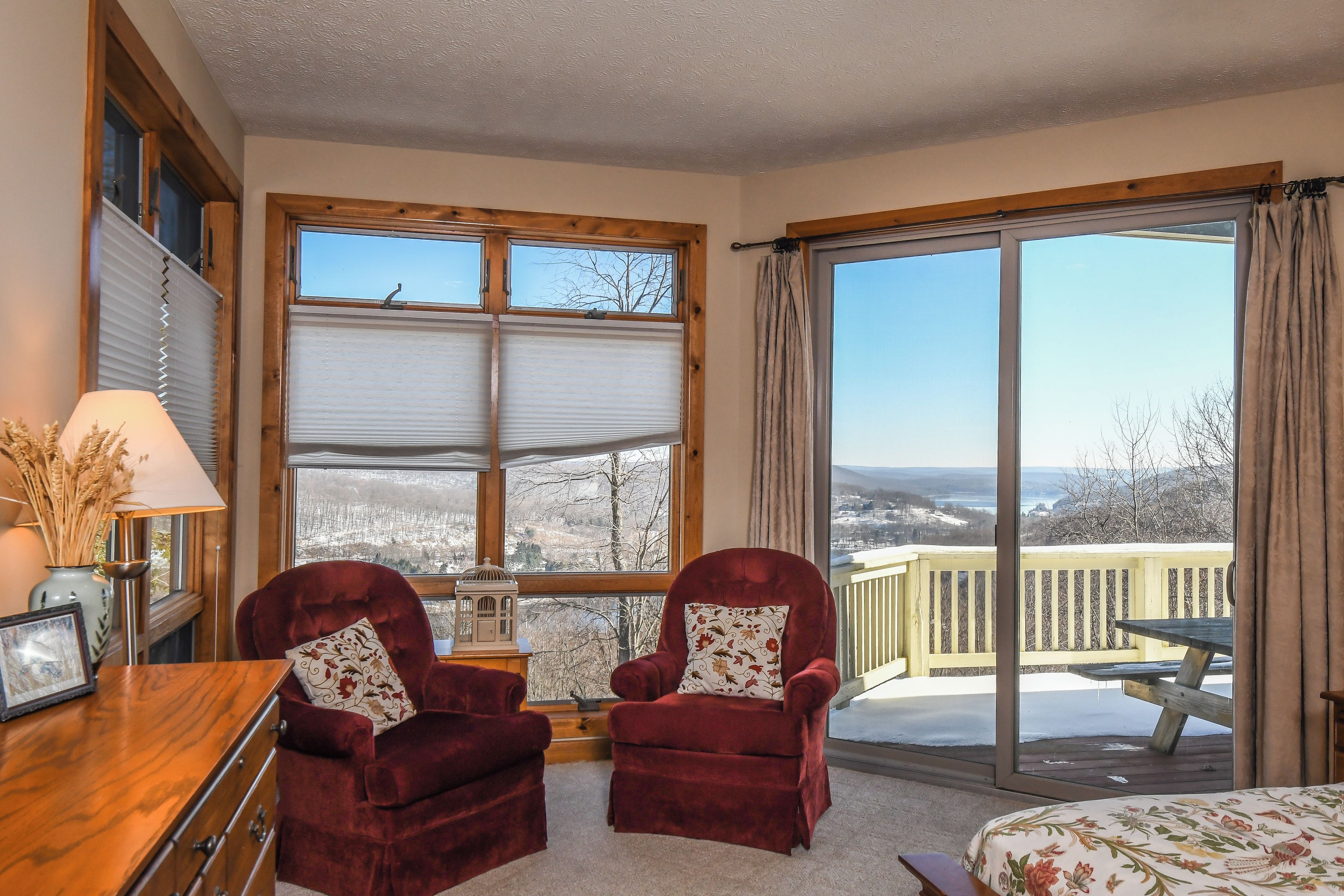 Ein Zimmer mit zwei roten Samtarmlehnen, einem Holztisch, einem großen Fenster mit Jalousien und Blick auf eine verschneite Landschaft durch eine Schiebeglastür.