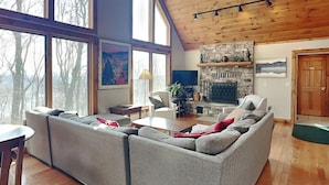 A spacious living room with a stone fireplace, large windows, and a high wooden ceiling. - One Ski Hill by Taylor Made Deep Creek (McHenry)
