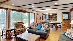 A spacious living room with a wooden coffee table, two sofas with floral cushions, a dining area with a wooden table and chairs, and large windows offering a view of the outdoors. - Our Golden Retreat by Taylor Made Deep Creek (Swanton)