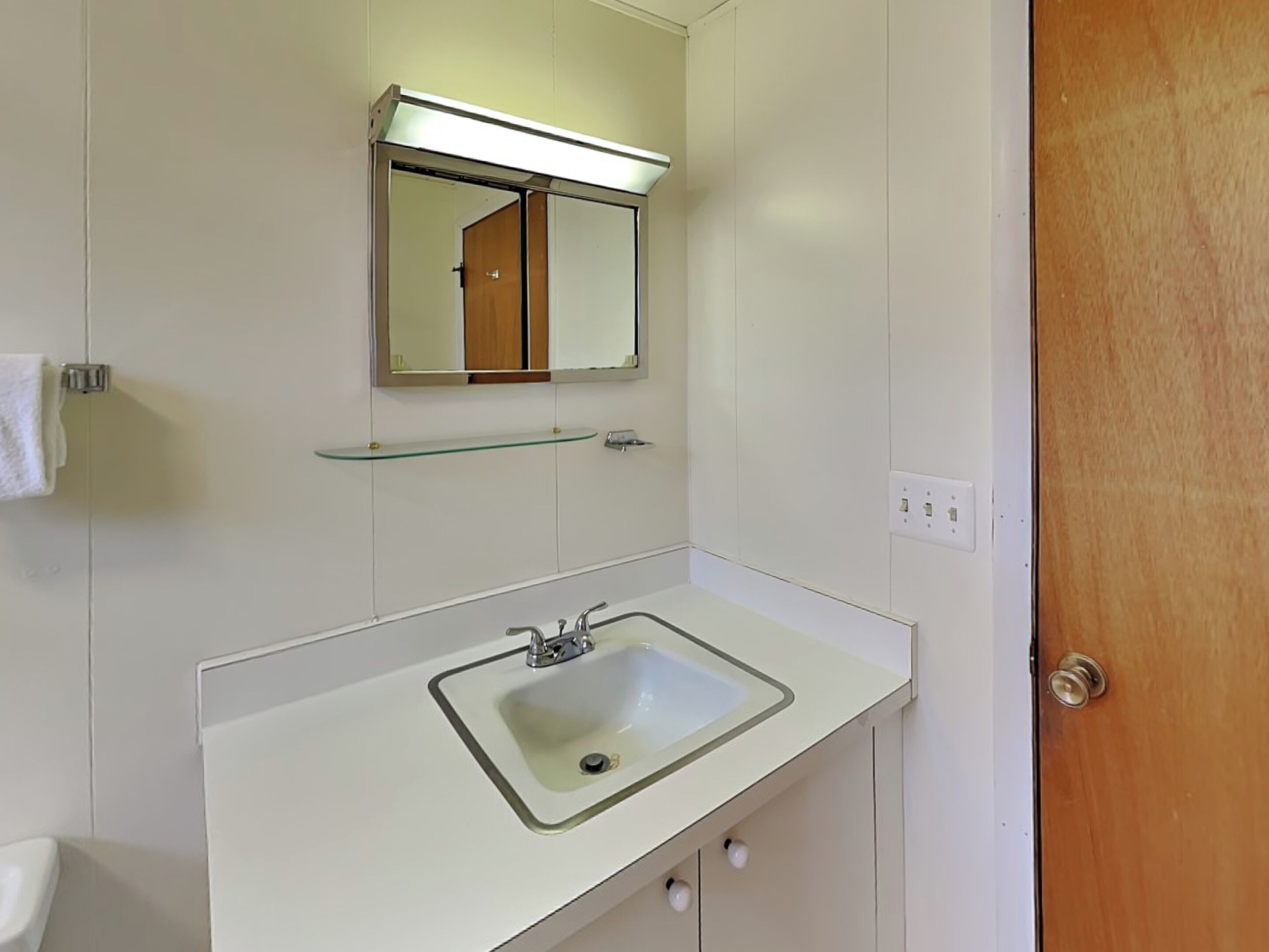 Un baño con un lavamanos blanco, un espejo encima y una repisa de vidrio.