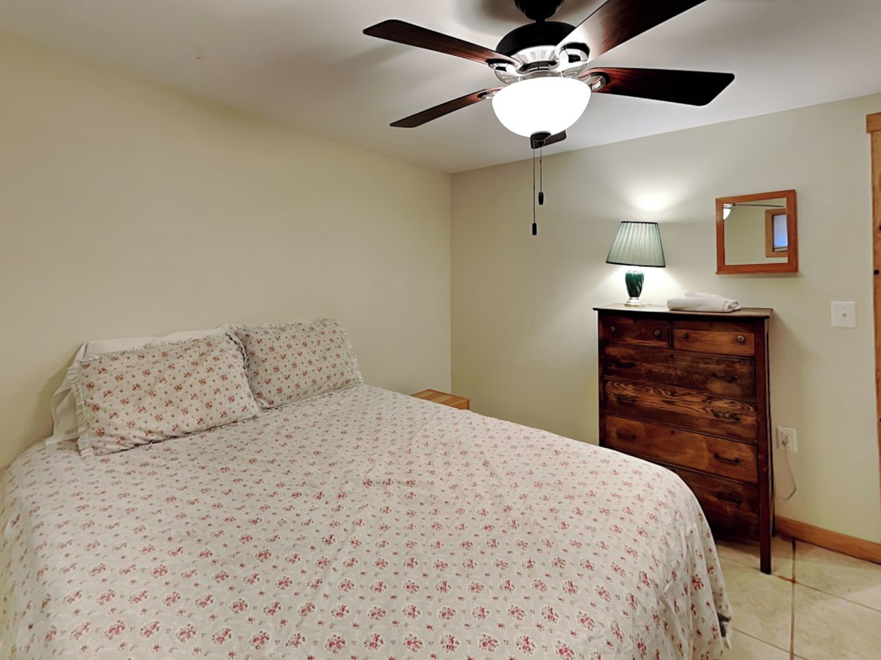 A bedroom with a bed, a wooden dresser, a ceiling fan, a lamp, and a mirror on the wall.