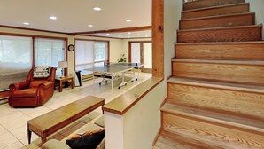 A spacious living room with a wooden staircase, a leather chair, a coffee table, and a ping pong table. - Our Golden Retreat by Taylor Made Deep Creek (Swanton)