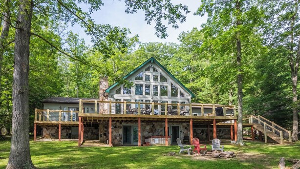 Une cabane à deux étages, avec des fondations en pierre, une terrasse en bois et un espace extérieur aménagé avec des sièges.