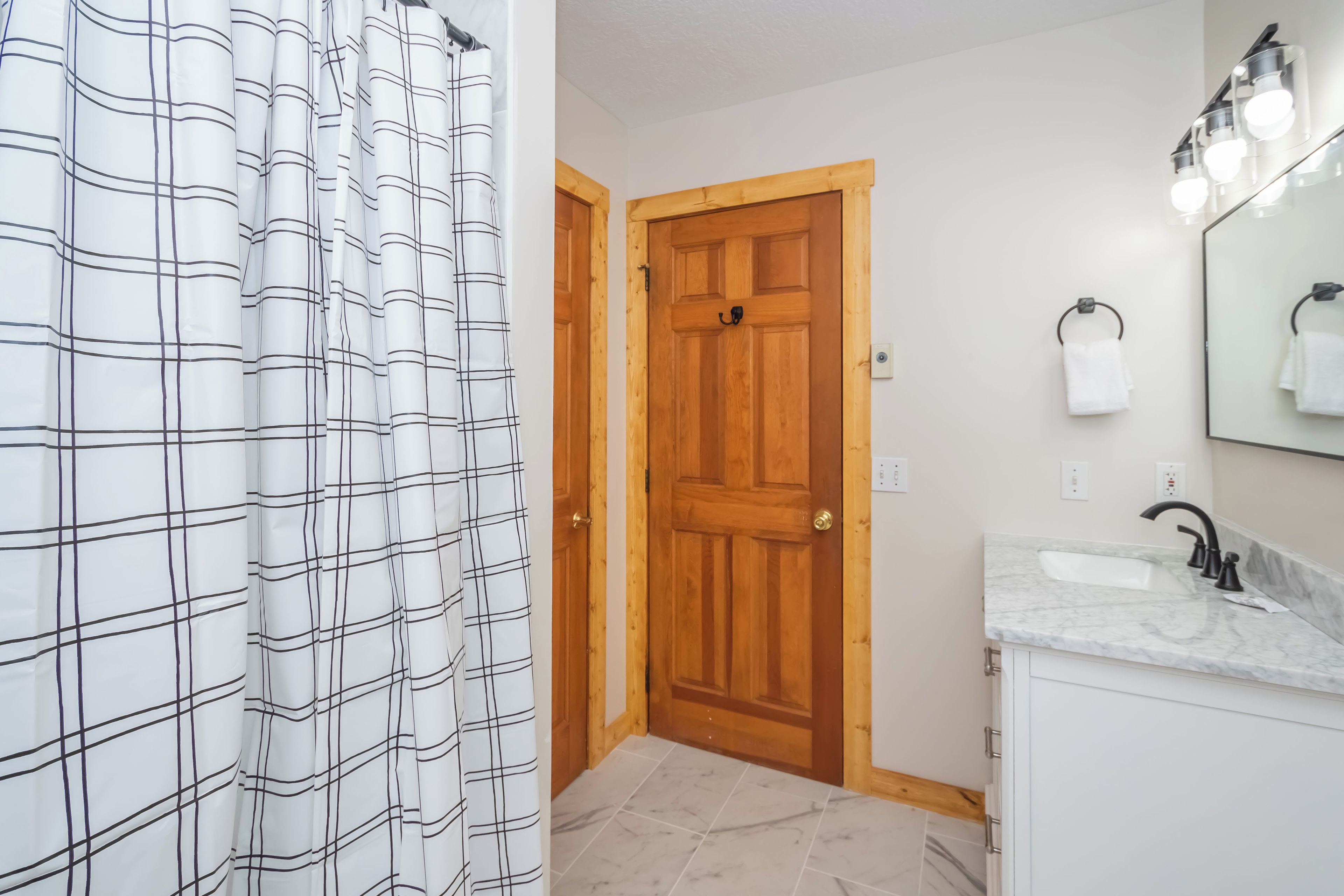 A bathroom with a wooden door, a white sink with a marble countertop, and a mirror with lights above it.