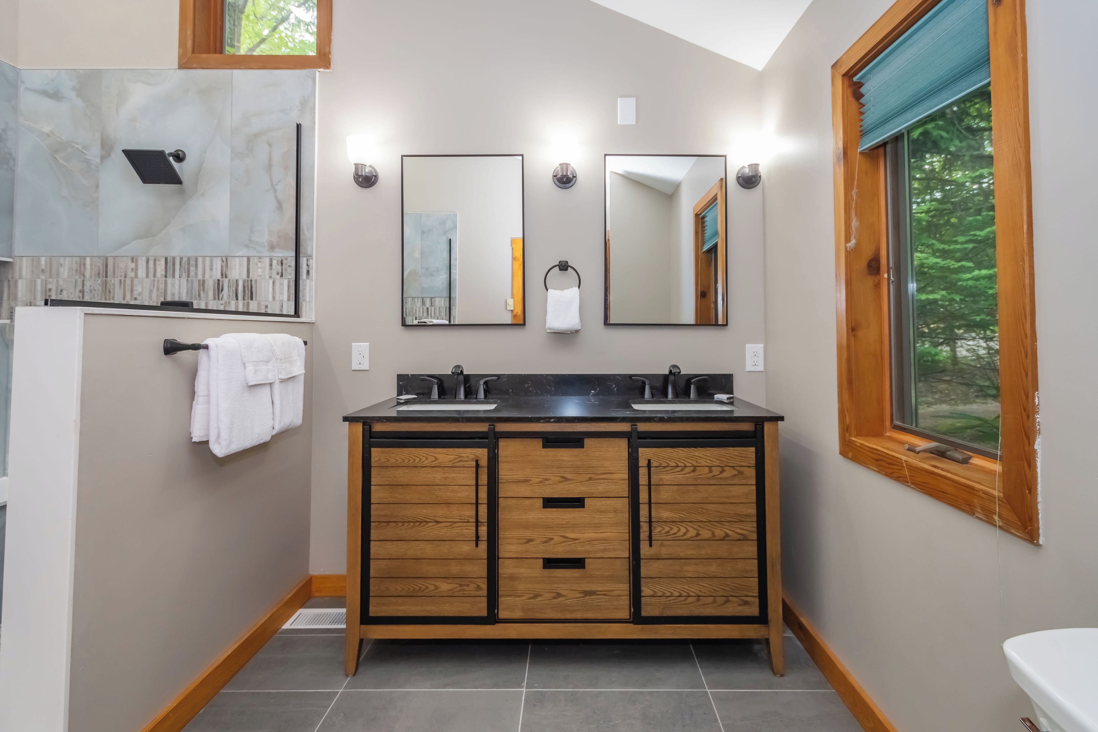 A modern bathroom with a wooden vanity, two sinks, a glass shower enclosure, and a window with a view of trees.