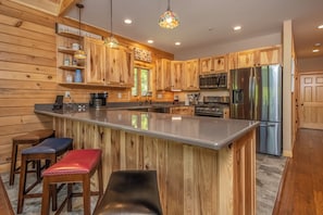 A modern kitchen with wooden cabinetry, a stainless steel refrigerator, and a bar area with stools. - Parhelion Chalet by Taylor Made Deep Creek (McHenry)