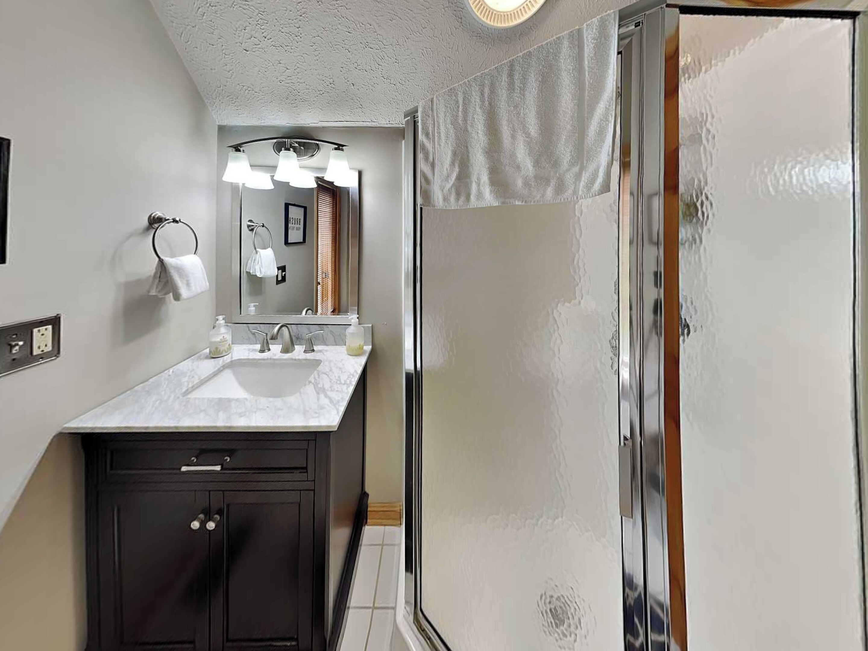 A modern bathroom with a marble sink, dark cabinets, a large mirror, and a glass shower enclosure.