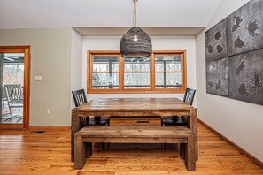 Un comedor con mesa de madera y bancos, ventanas amplias y una lámpara colgante.