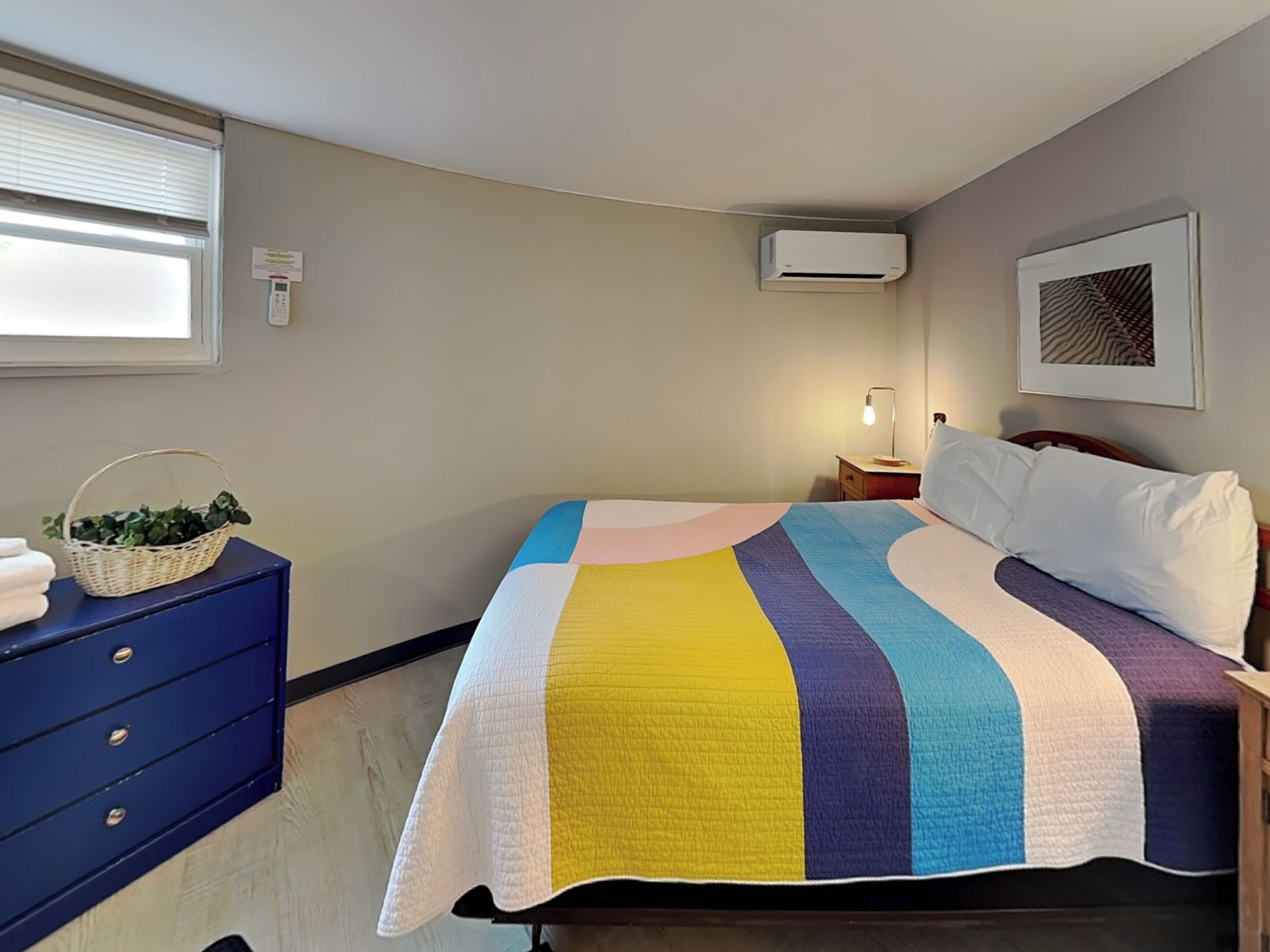 Une chambre à coucher comprenant un lit, une commode, une lampe, un climatiseur fixé au mur et une fenêtre avec des stores.