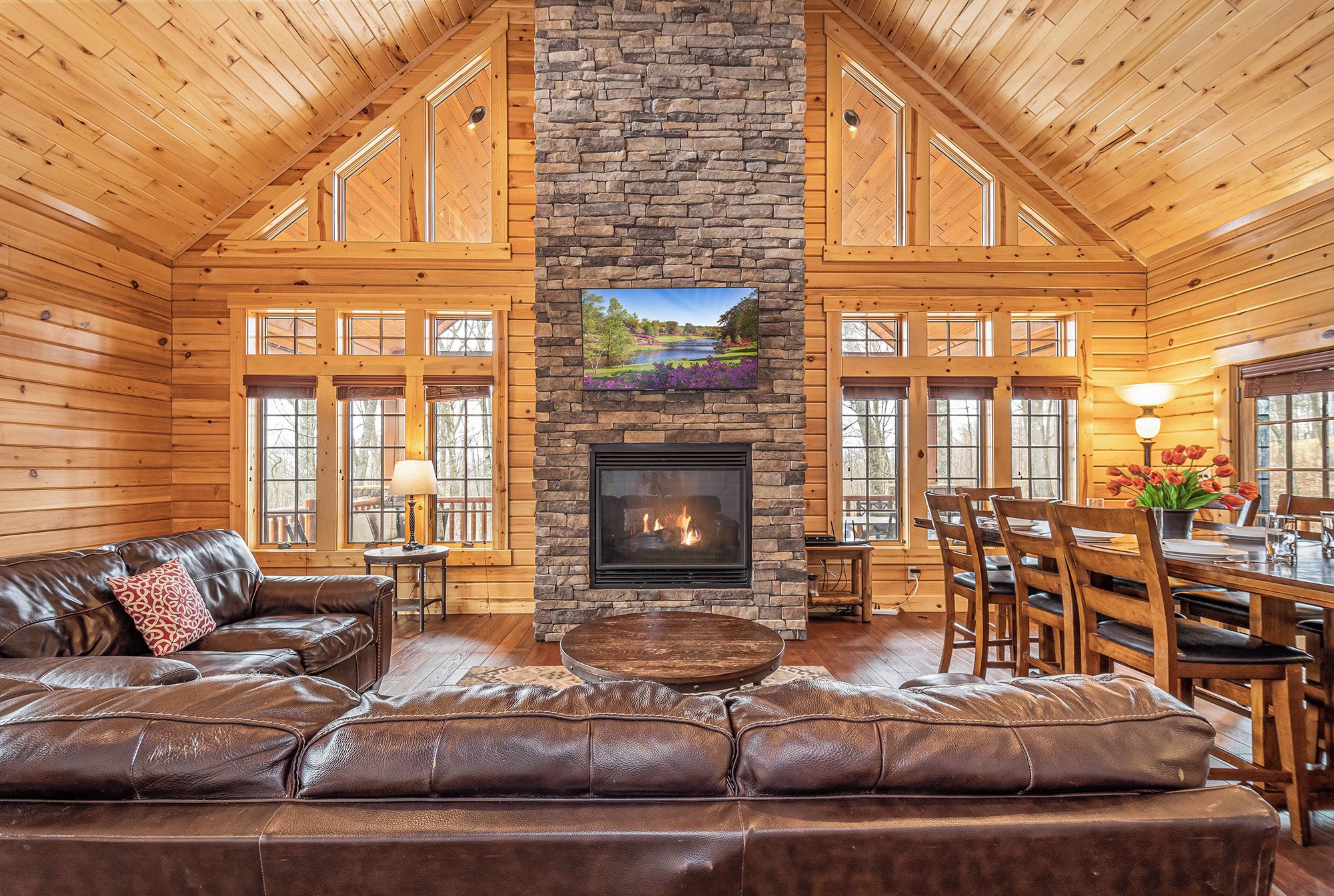 A log cabin interior with a stone fireplace, leather sofa, dining area, and large windows.