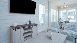 A modern living room with a flat-screen TV mounted on the wall, a wooden entertainment center, patterned seating, and large windows. - Sandy of Panama City Beach - Ocean View, Heated Pool, Steps to Beach, Sleeps 18 (Panama City Beach)