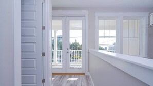 A narrow hallway with white walls and wooden flooring leading to a balcony with white railings and a view of buildings and trees. - Sandy of Panama City Beach - Ocean View, Heated Pool, Steps to Beach, Sleeps 18 (Panama City Beach)