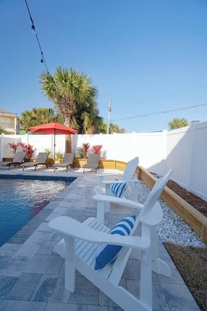 Un espace aménagé au bord de la piscine, avec des chaises longues blanches, un parasol rouge et un palmier.